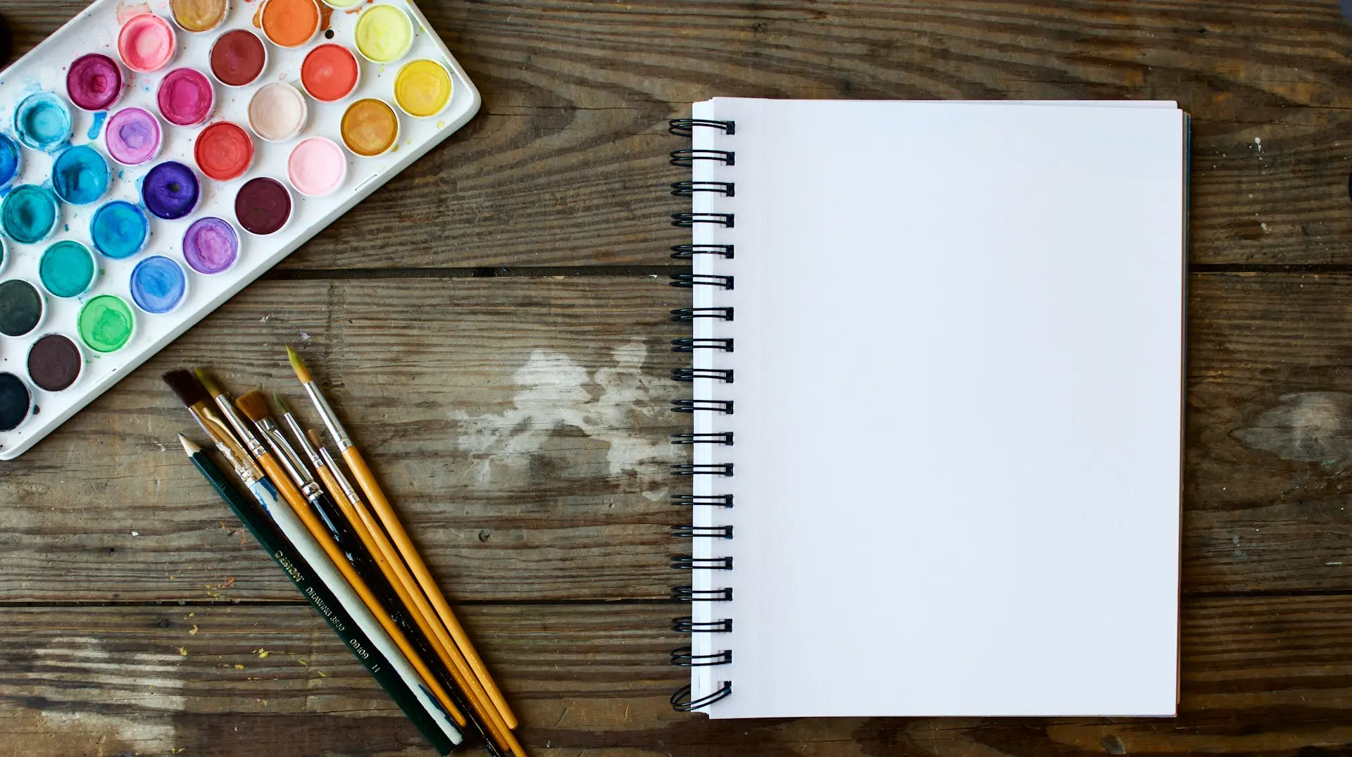 paint brushes next to drawing book and water color palette
